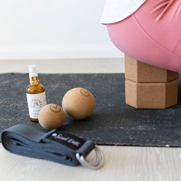 Woman sat on cork yoga blocks laid on black jute eco-yoga mat, with meditation spray, cork balls and a yoga strap next to her, taking time out in the office